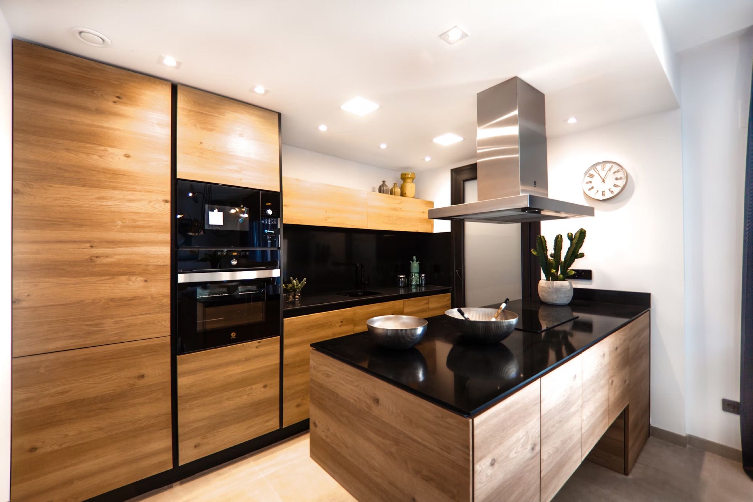 A kitchen with oak-effect tall units, a black island worktop and a stainless steel hood